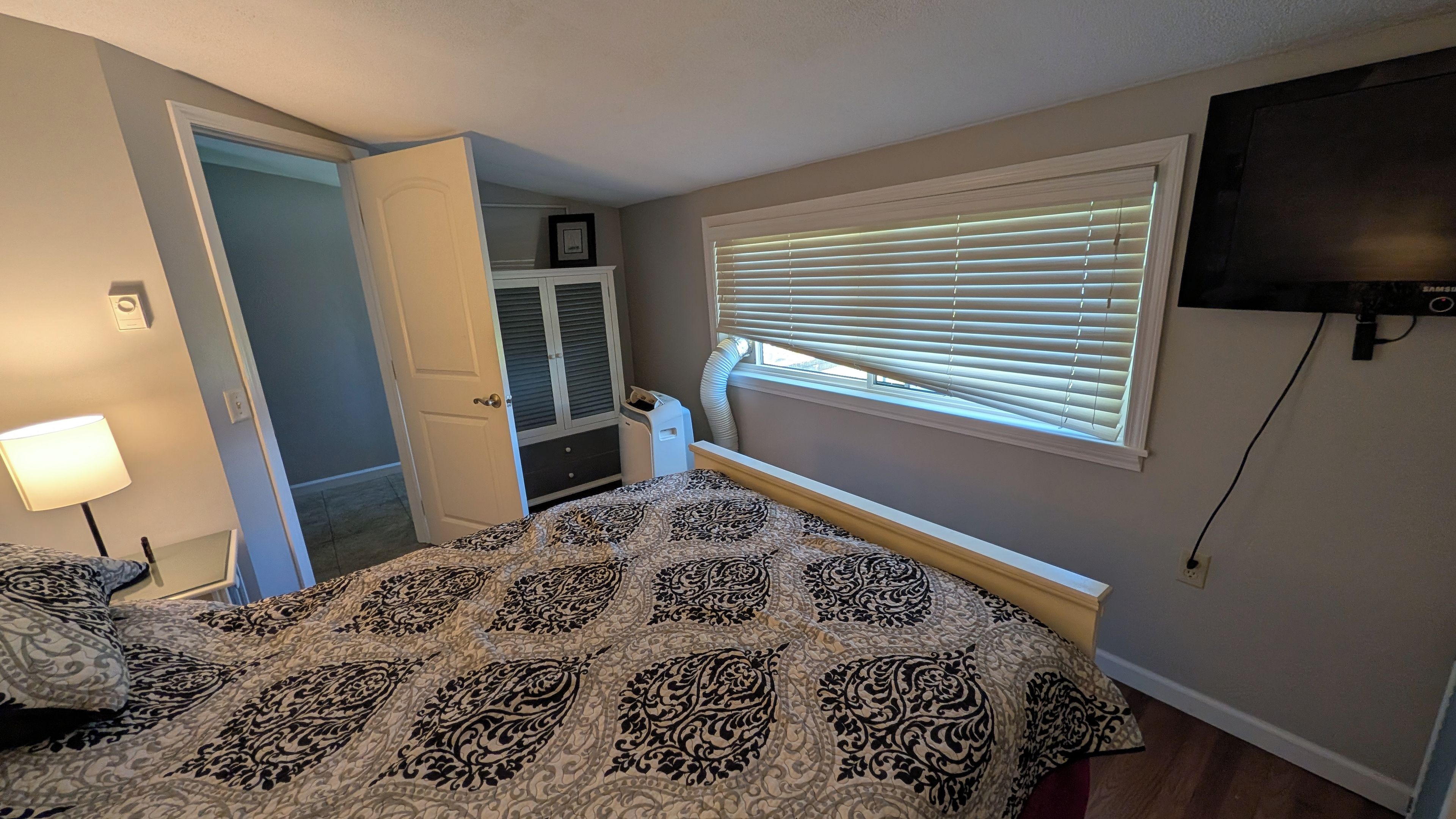 Une chambre à coucher avec un lit, un téléviseur fixé au mur, une fenêtre avec des stores, une porte et un petit radiateur.