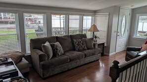 Ein Wohnzimmer mit einem dunkelbraunen Sofa, gemusterten Kissen, einem Beistelltisch mit Lampe und großen Fenstern mit Jalousien.