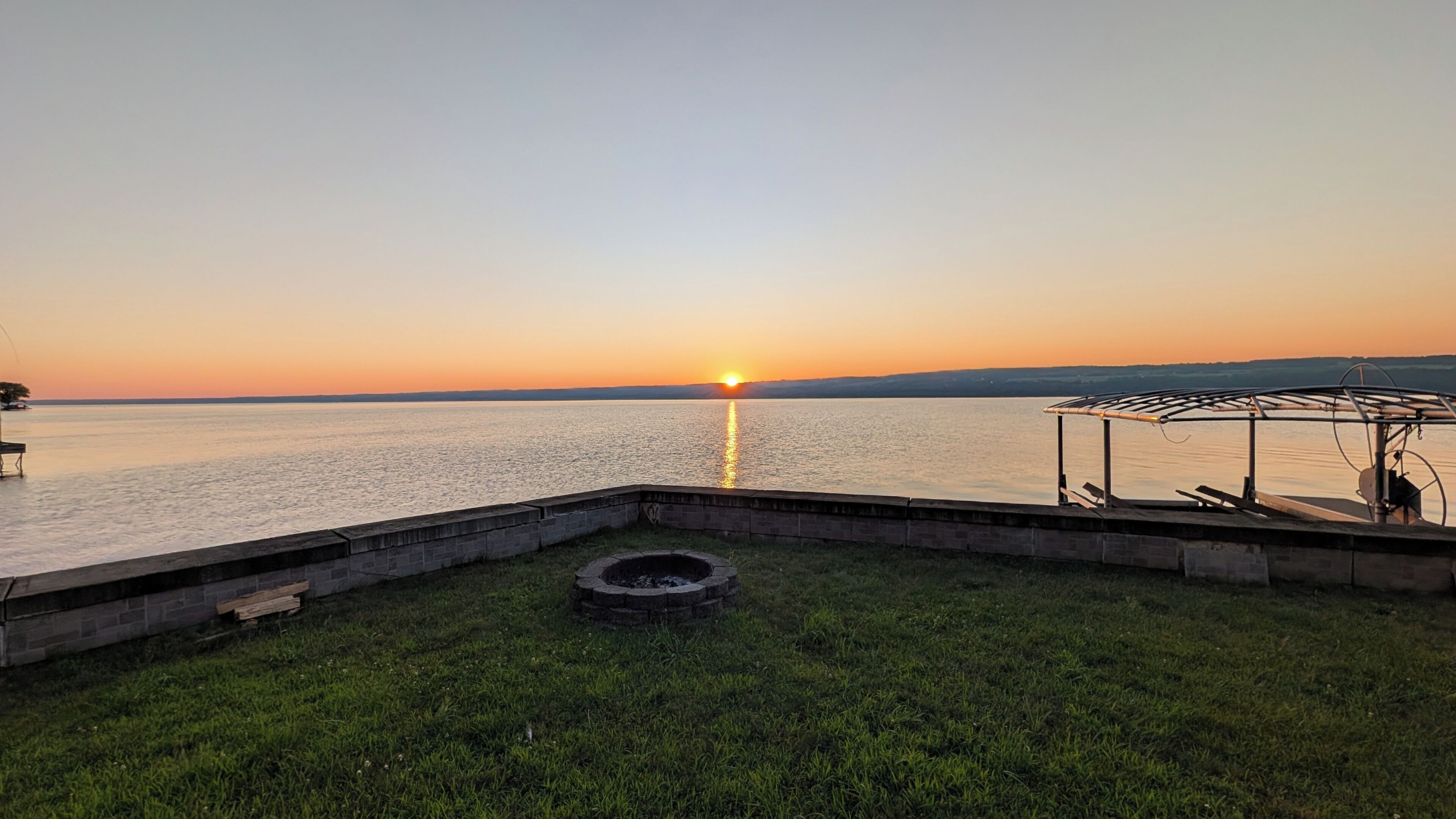 Une vue sur le coucher de soleil au-dessus d’un lac paisible, avec une zone herbeuse et une structure abritée.