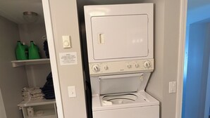 A laundry room with a stacked washer and dryer, a shelf with folded towels, and a door leading to another room.