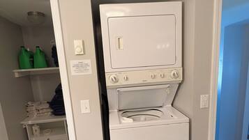 A laundry room with a stacked washer and dryer, a shelf with folded towels, and a door leading to another room.