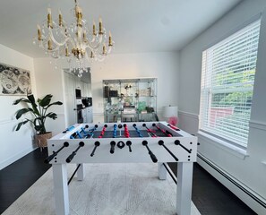A room with a foosball table, a chandelier, a large window with blinds, and a plant.