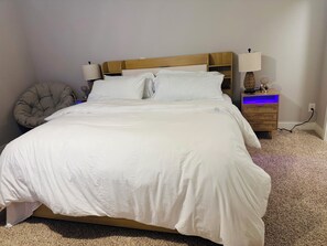 A neatly made bed with white bedding, a wooden headboard, a bedside lamp, a small wooden nightstand with a lamp, and a chair.