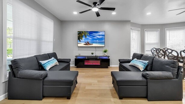 A modern living room with two black leather sofas, a ceiling fan, a flat-screen TV, and a fireplace.