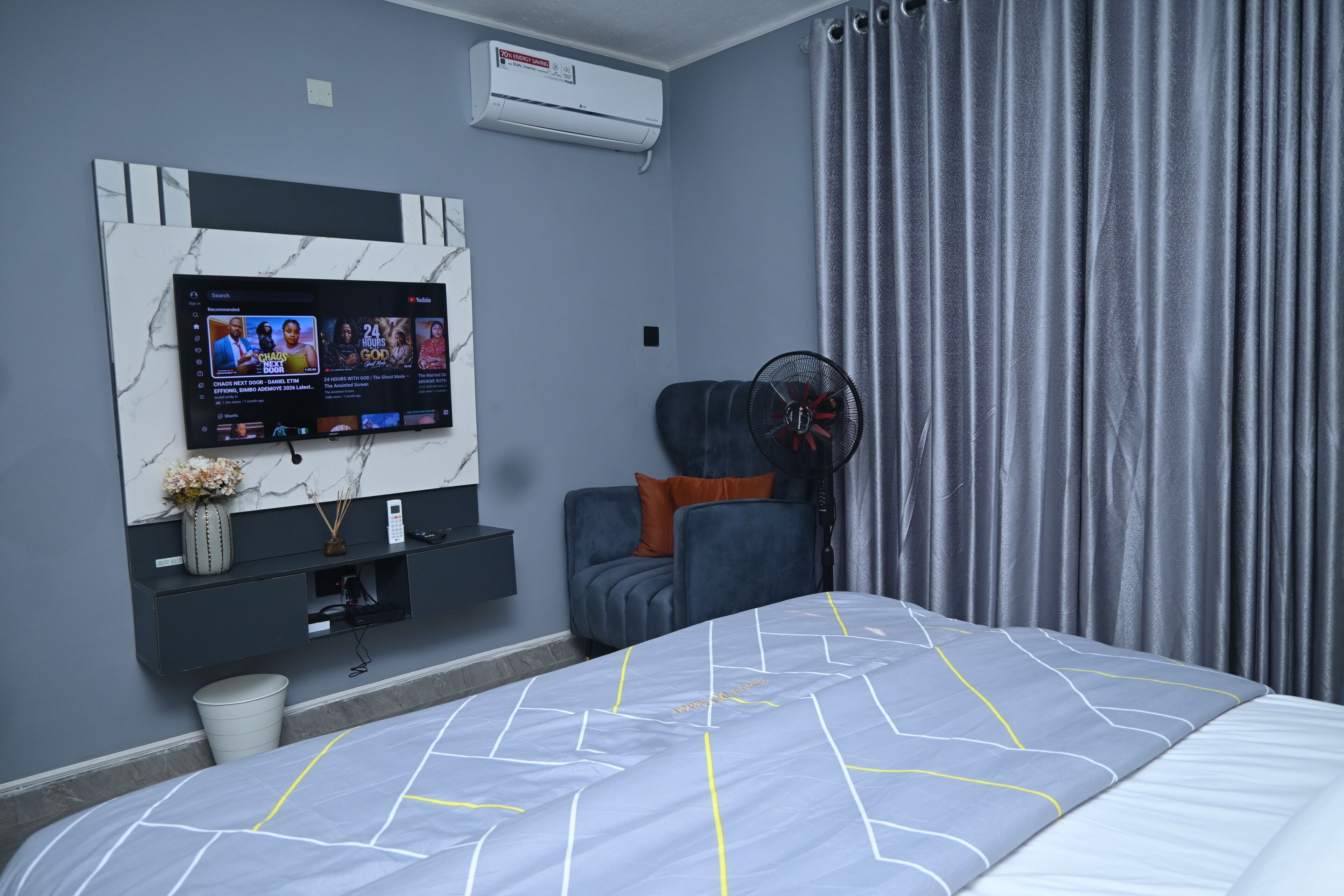 Una camera d'albergo moderna con un letto, una TV a parete, una sedia, una ventola e una tenda.