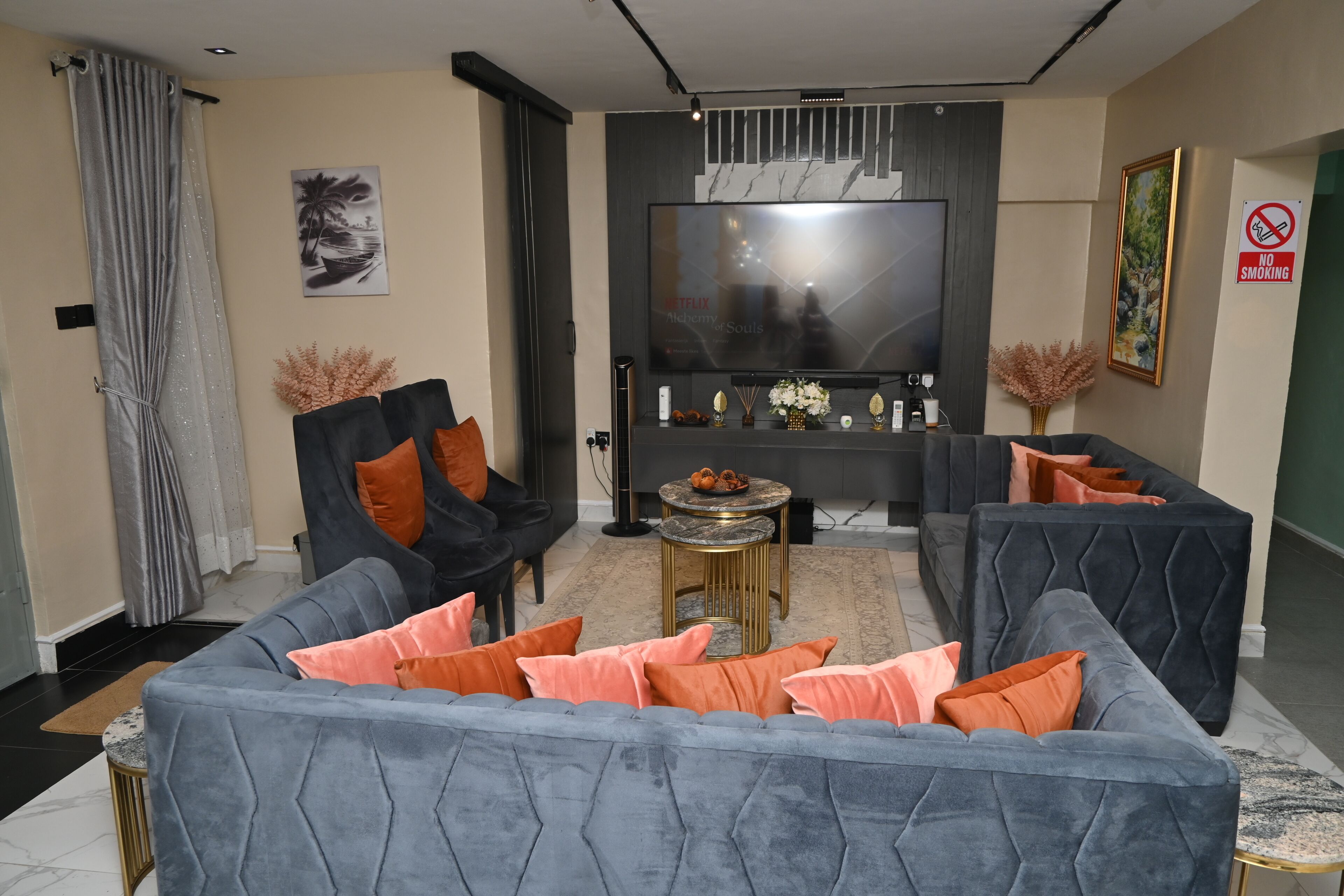 Un salon moderne avec un canapé gris, des coussins orange, un téléviseur à écran plat et un panneau indiquant « Il est interdit de fumer ».