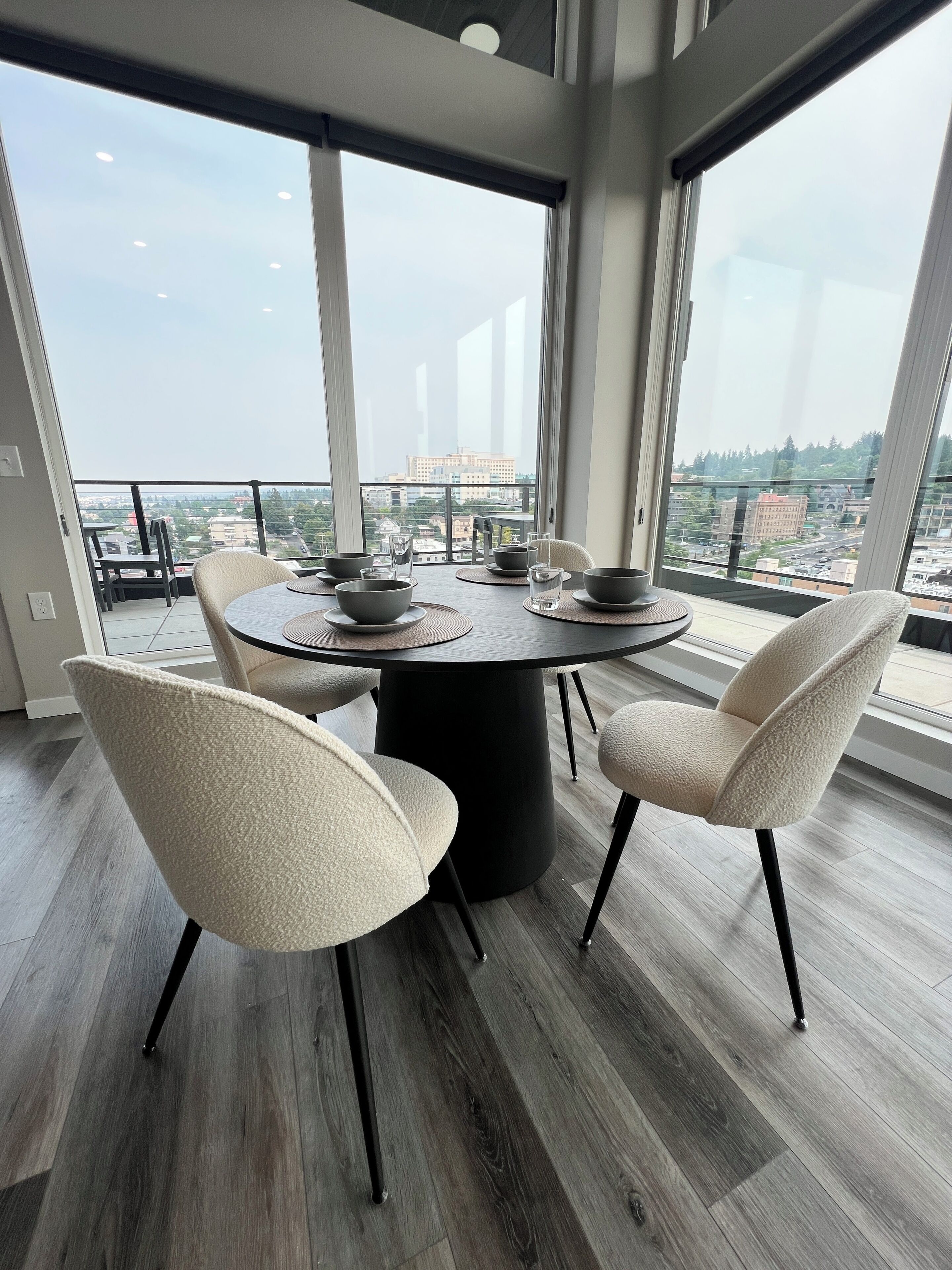 A modern dining area with a round table, four chairs, and a view of the cityscape through large windows.