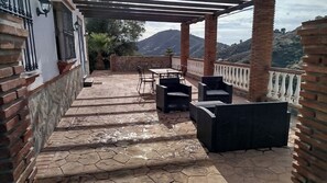 A covered outdoor patio with stone flooring, wicker furniture, and a view of hills. - Casa Miranda Cómpeta by Ruralidays (Málaga)