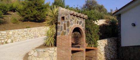 A stone and brick outdoor oven structure with a small opening, surrounded by a stone wall and vegetation.