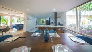 A dining area with a wooden table set for four, a large blue vase centerpiece, and a view of a garden through large windows. - The Garden Mansion | 11 Bedrooms with Club Lounge•Jacuzzi•Olympic Pool•Maid•AC (Medellín)