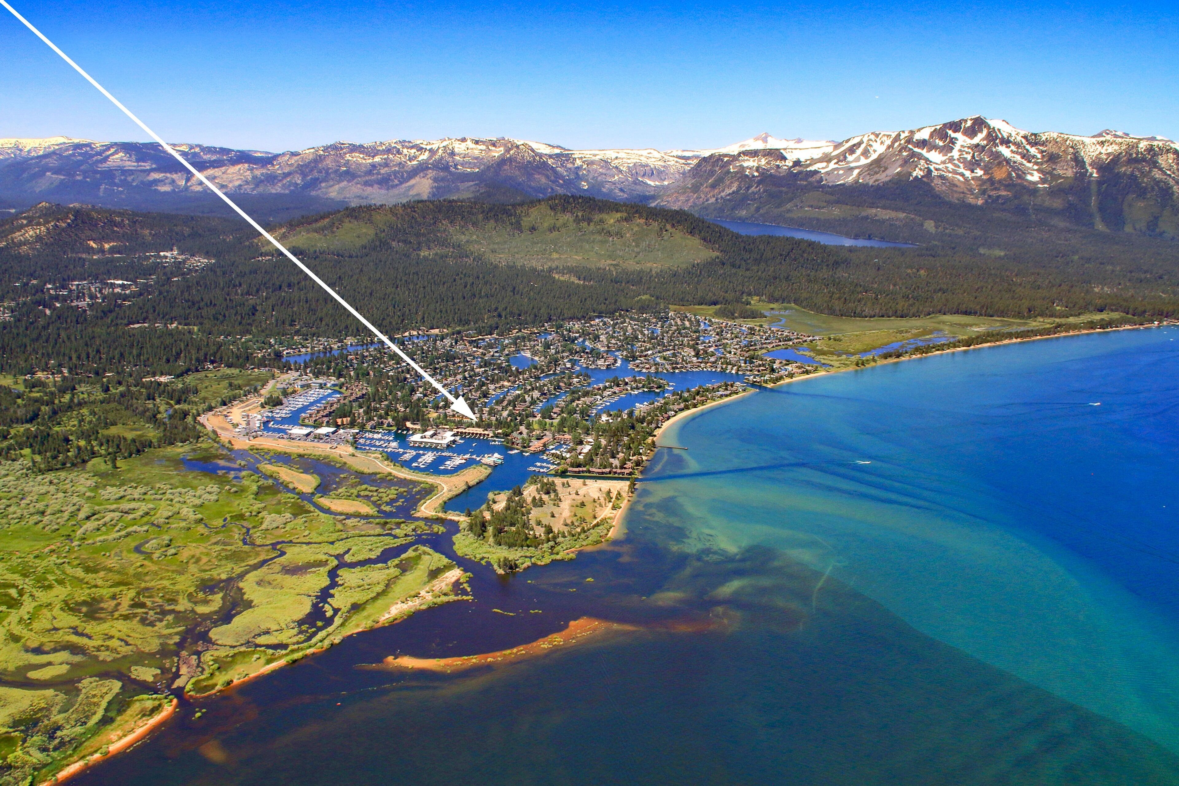 An aerial view of a coastal town with a marina, clear blue waters, and a backdrop of snow-capped mountains.
