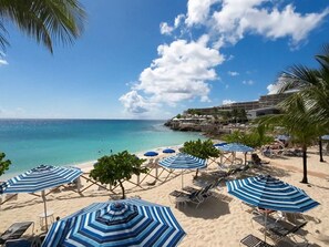 A beach with sun loungers and umbrellas, a clear sky, and a building in the background. - Prime location in Lowlands, Sint Maarten (Maho Bay)