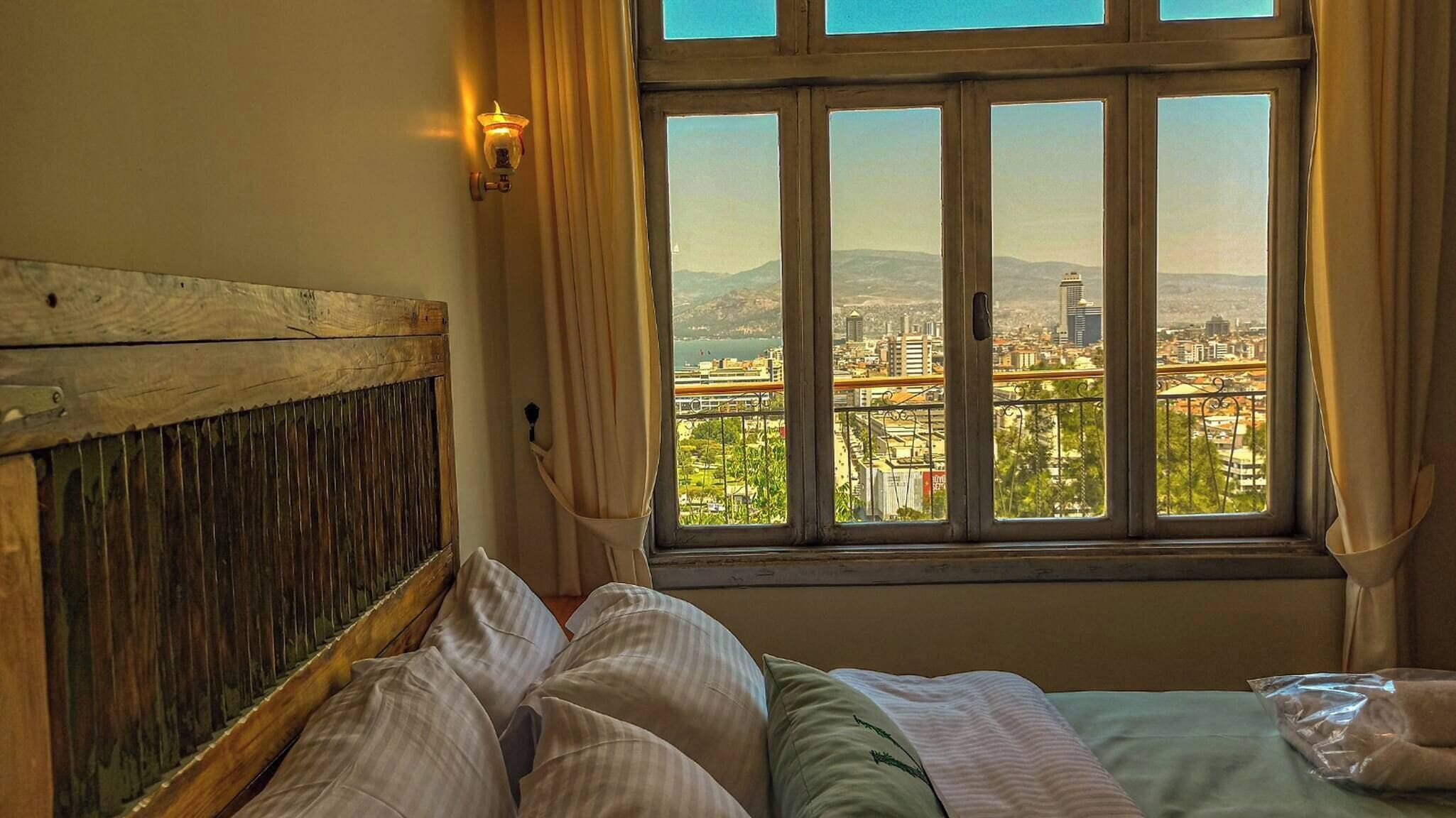 Une chambre avec un lit, une fenêtre donnant sur un paysage urbain et un luminaire fixé au mur.