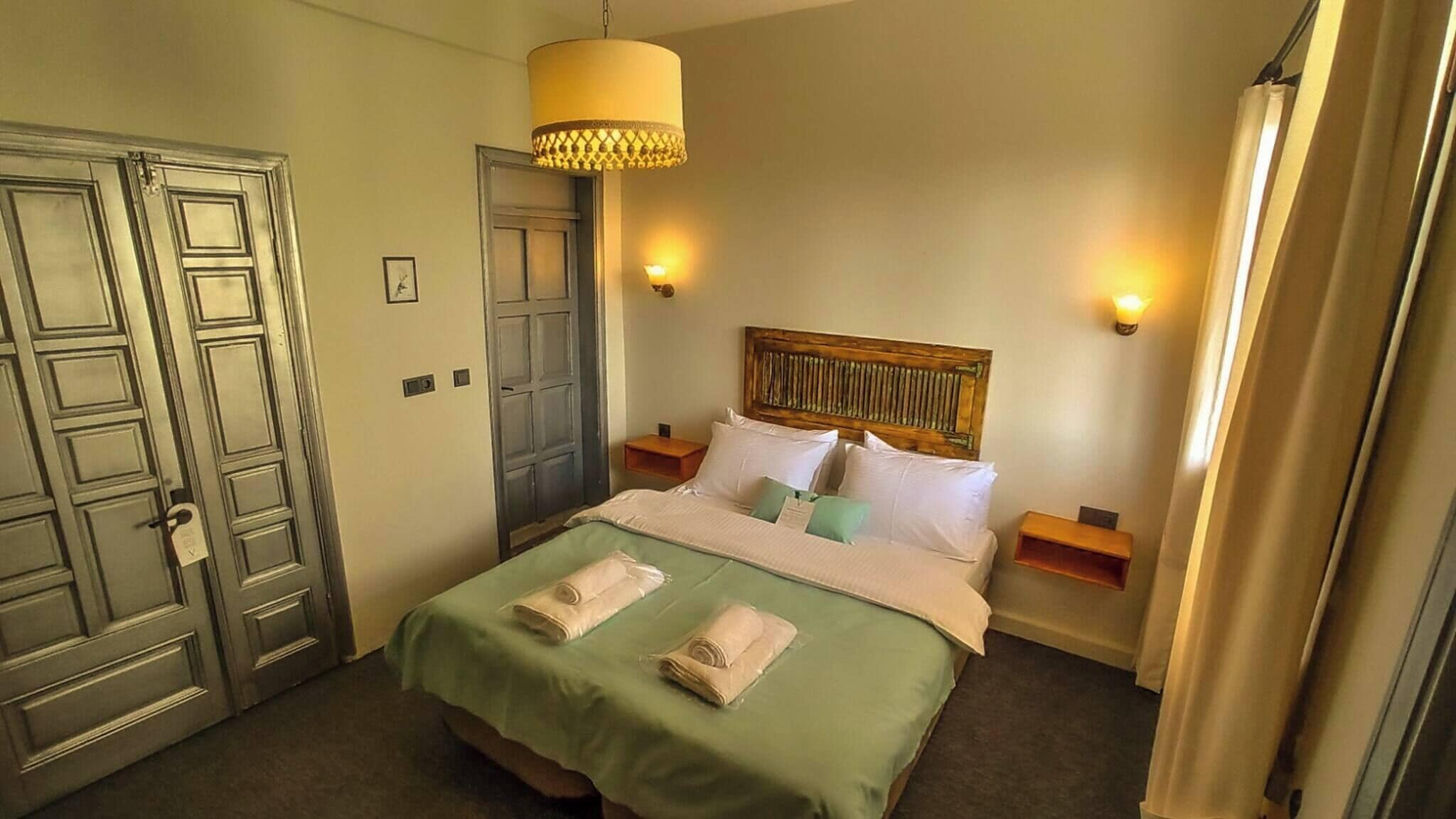 Une chambre d’hôtel comprenant un lit, deux tables de chevet, une tête de lit en bois, un luminaire suspendu et une porte.