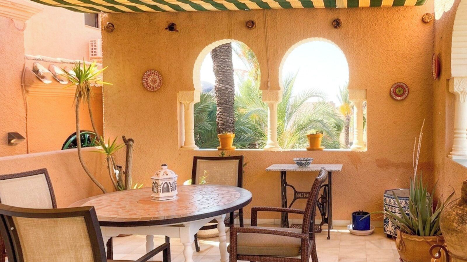 A cozy outdoor seating area with a round table and chairs, a small table with a bowl, and a window with a view of palm trees.