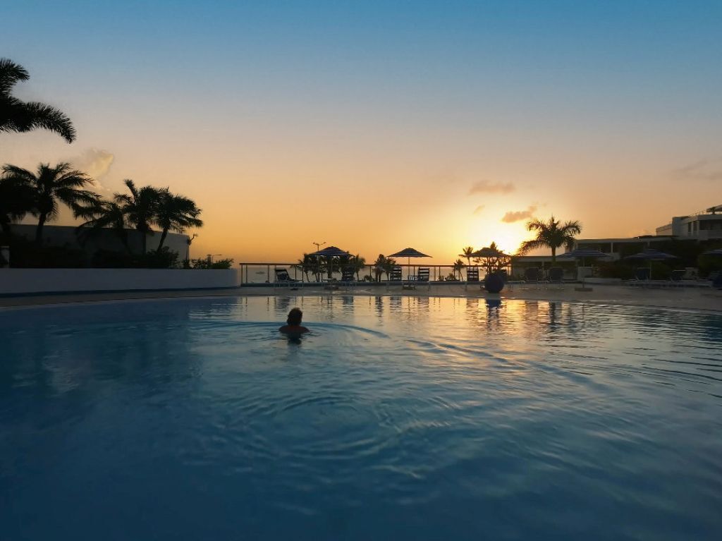 A swimming pool with a sunset in the background, palm trees, and a building with a roofed area.