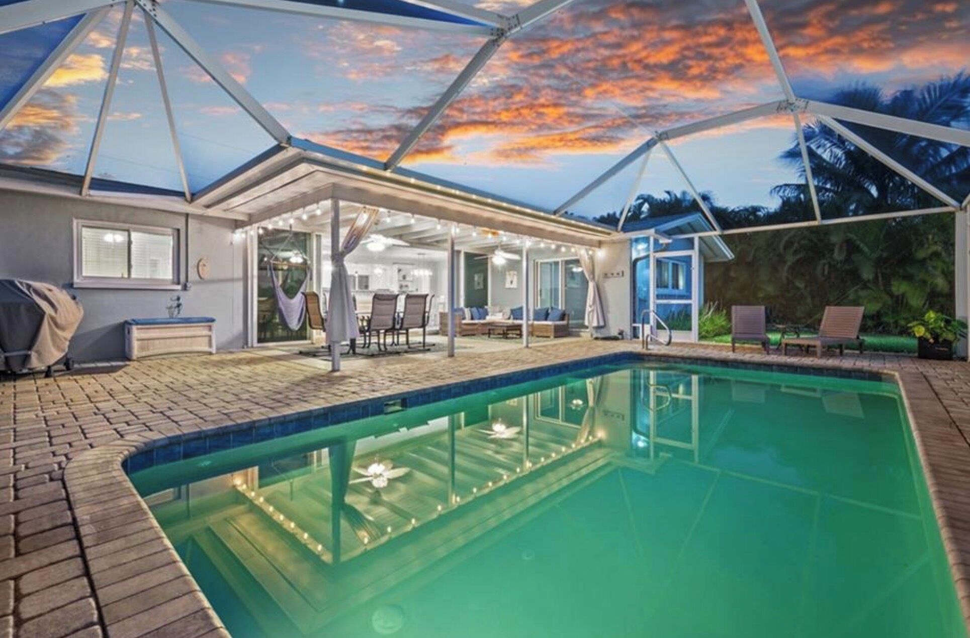 A swimming pool with a covered patio area, a dining table, and chairs.