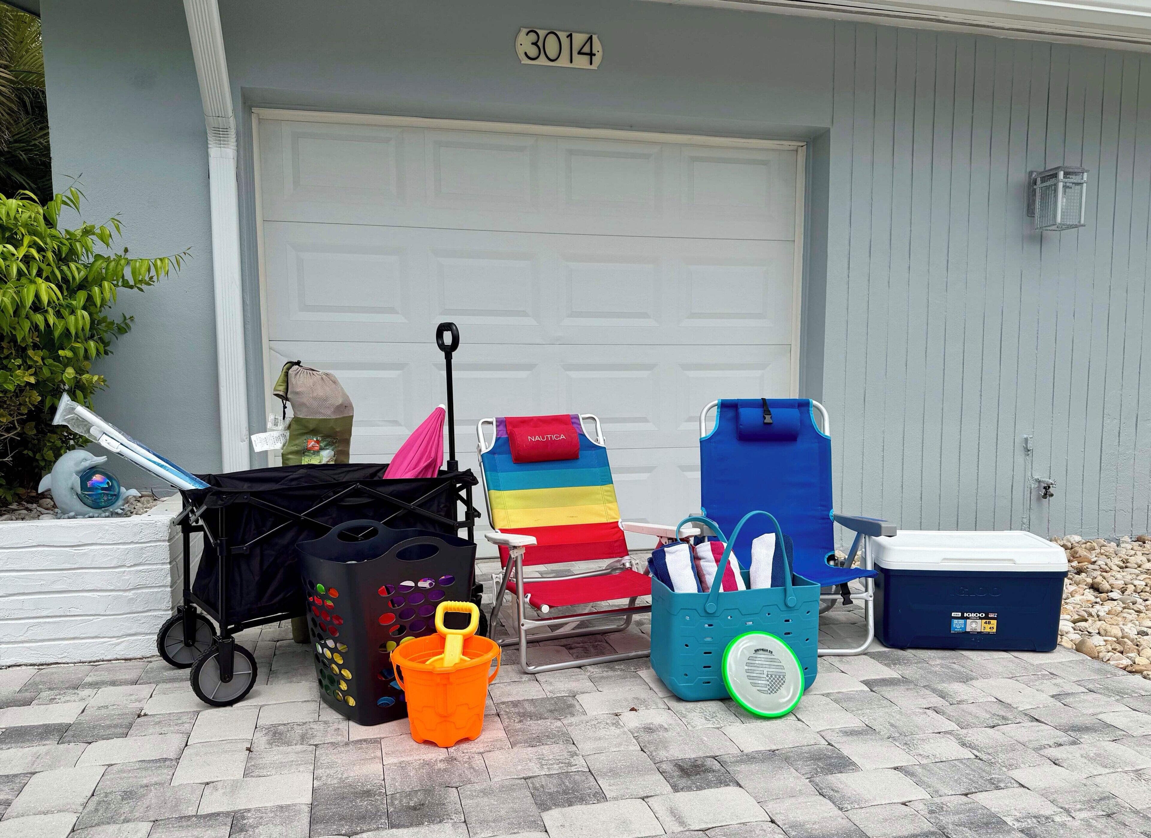 A garage door with the number 3014, a beach chair, a colorful beach umbrella, a black trash bin, a blue cooler, and a small orange bucket.