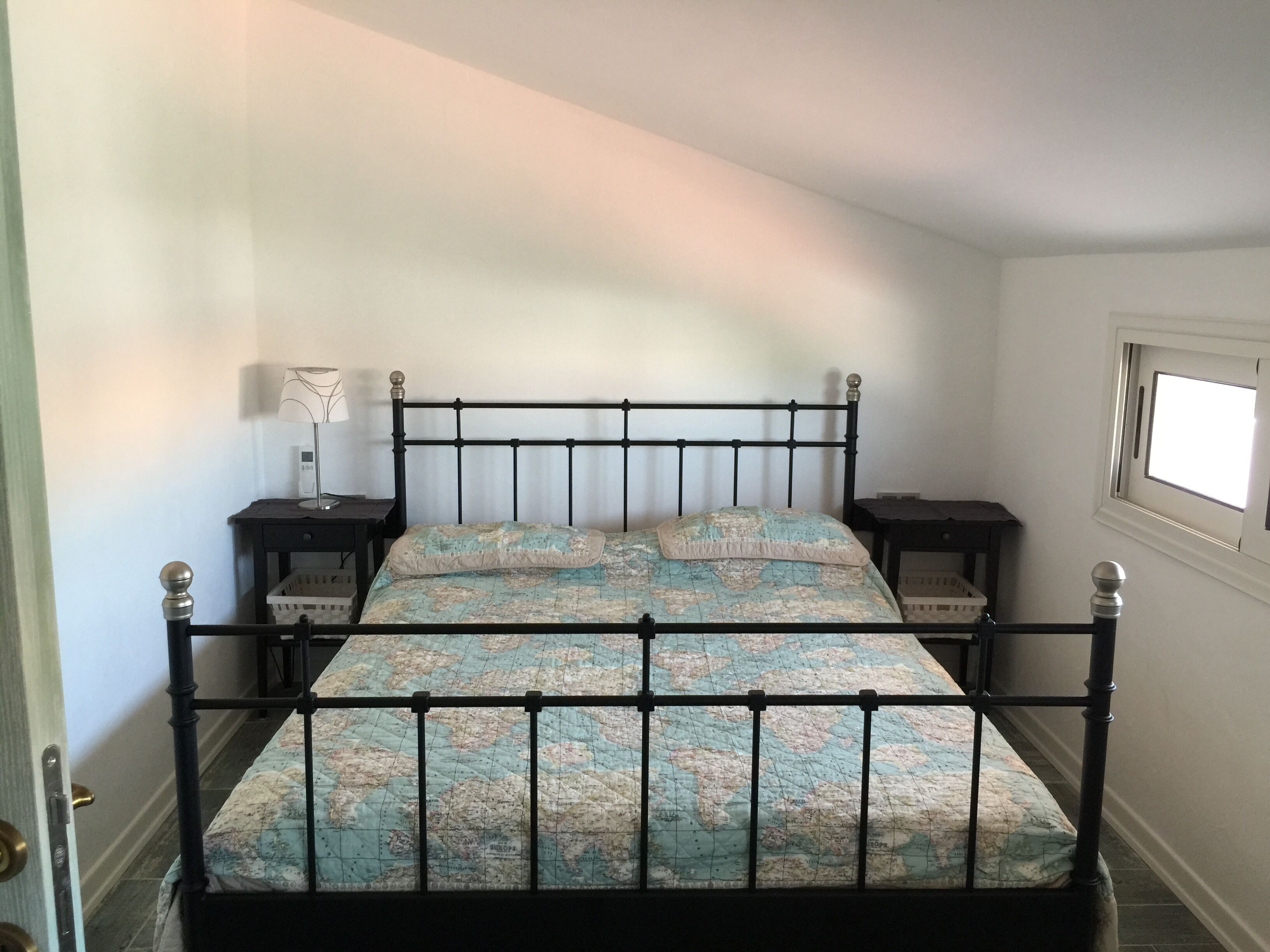 Una cama con estructura metálica, una colcha estampada con motivos de mapa, dos mesitas de noche, una lámpara y una ventana con vistas.