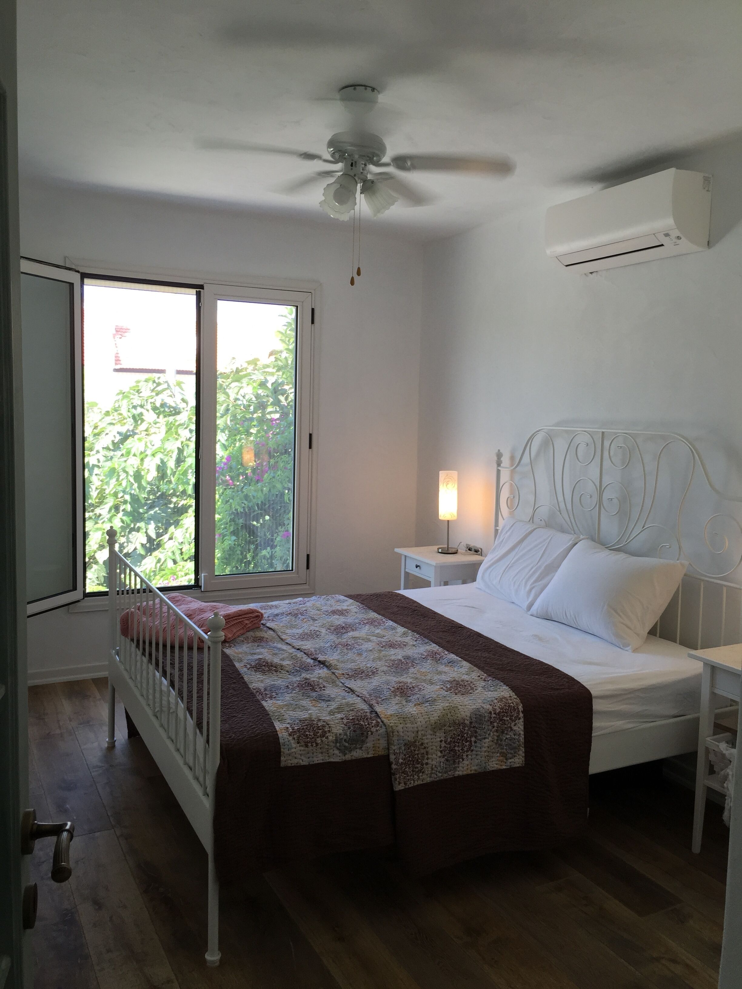 A bedroom with a bed, a nightstand, a lamp, a ceiling fan, and a window with a view of greenery.