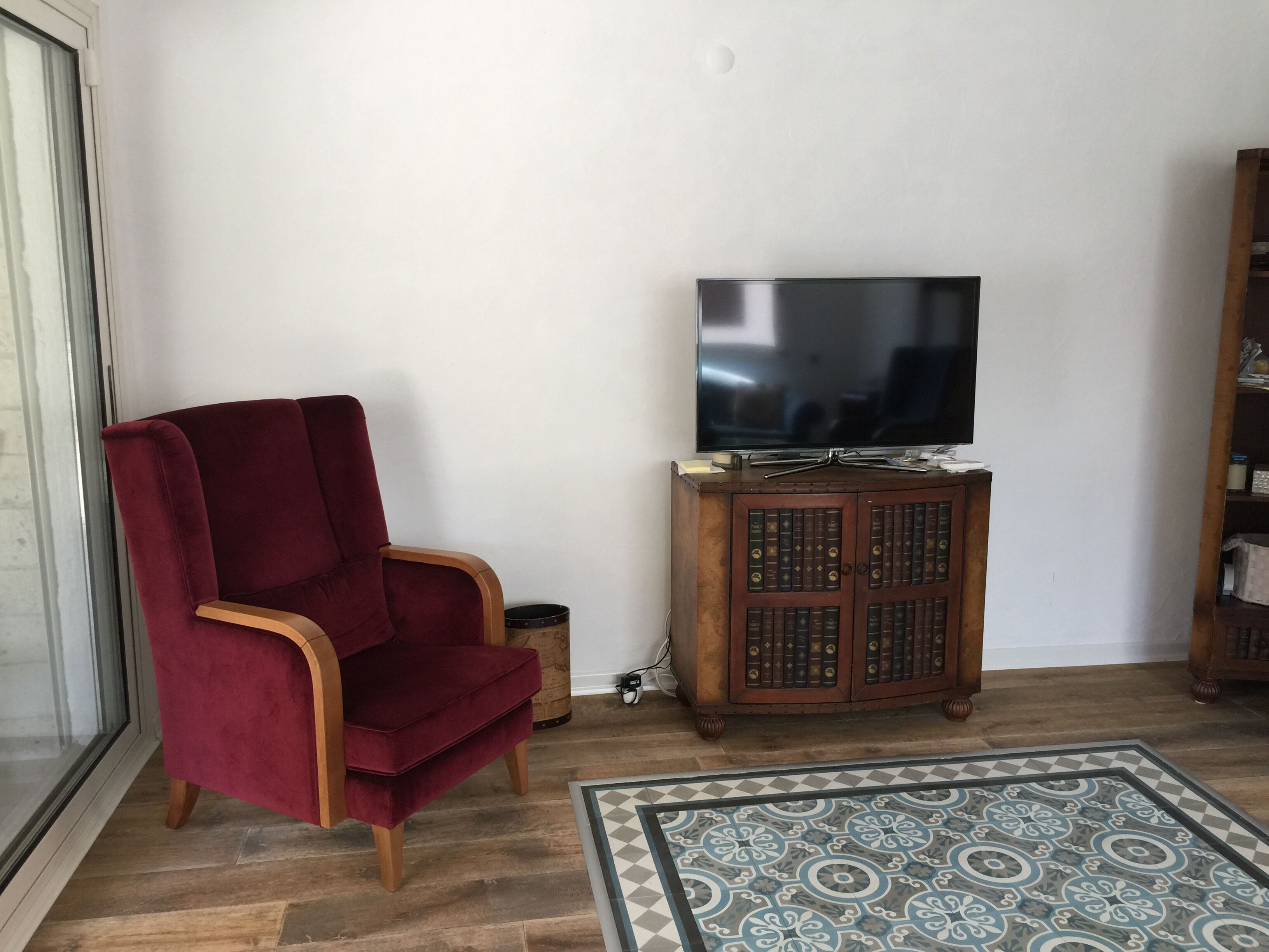 Un salon avec un fauteuil rouge, un meuble en bois sur lequel est posée une télévision, et un tapis à motifs.