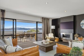 A modern living room with a fireplace, leather sofas, a dining area with chairs, and a large window overlooking a scenic view. - Harvey Heights Retreat (Taupō)