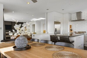 A modern kitchen with white cabinetry, a stainless steel range hood, and a dining area with a wooden table and woven placemats. - Harvey Heights Retreat (Taupō)