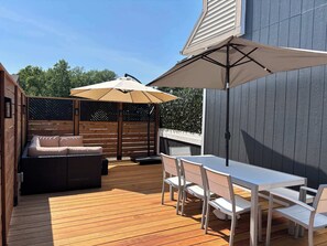 A wooden deck with a white table and chairs, a beige sofa, and two umbrellas providing shade.