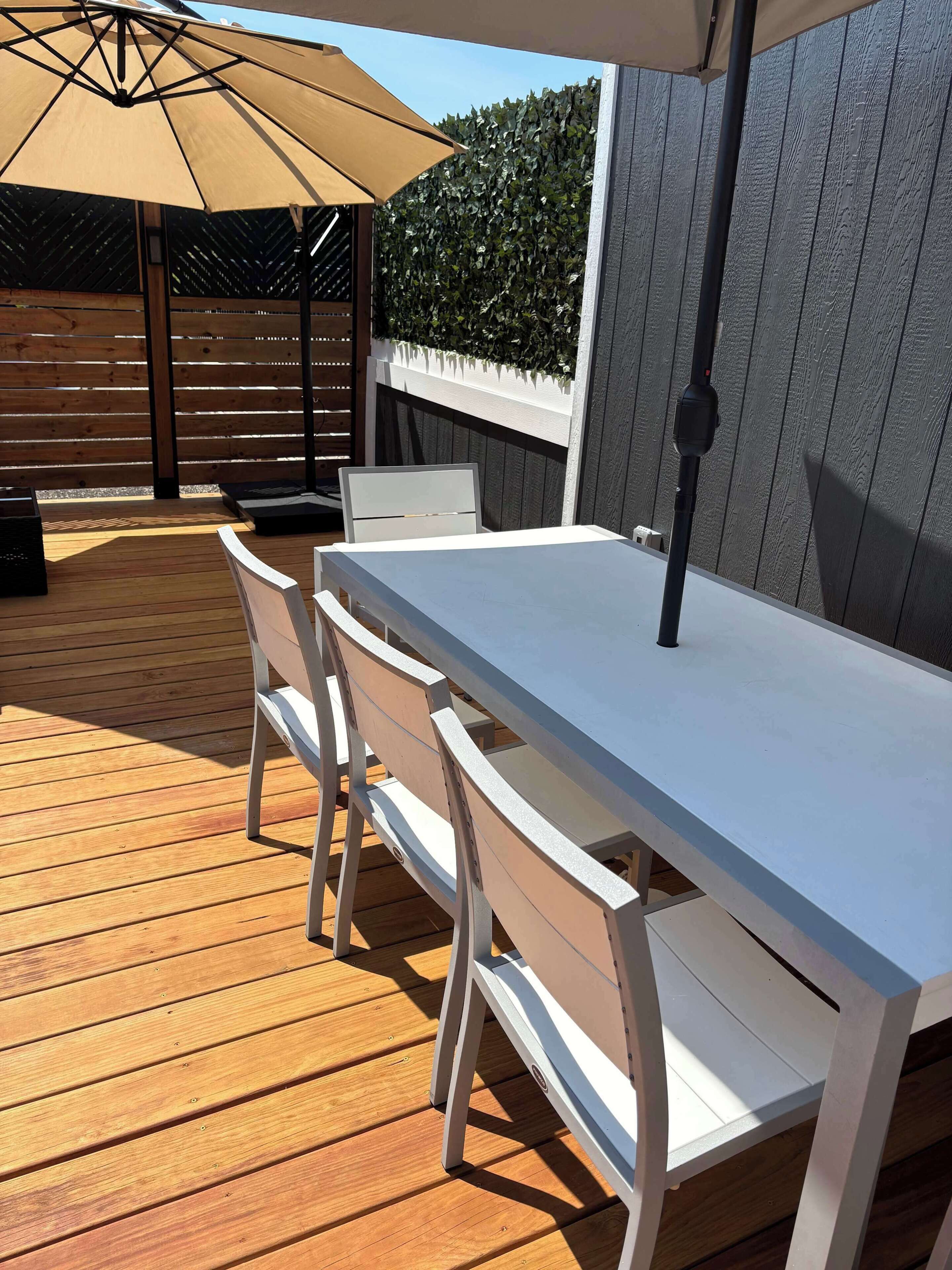 An outdoor patio with a white table and chairs, a beige umbrella, and a wooden deck.