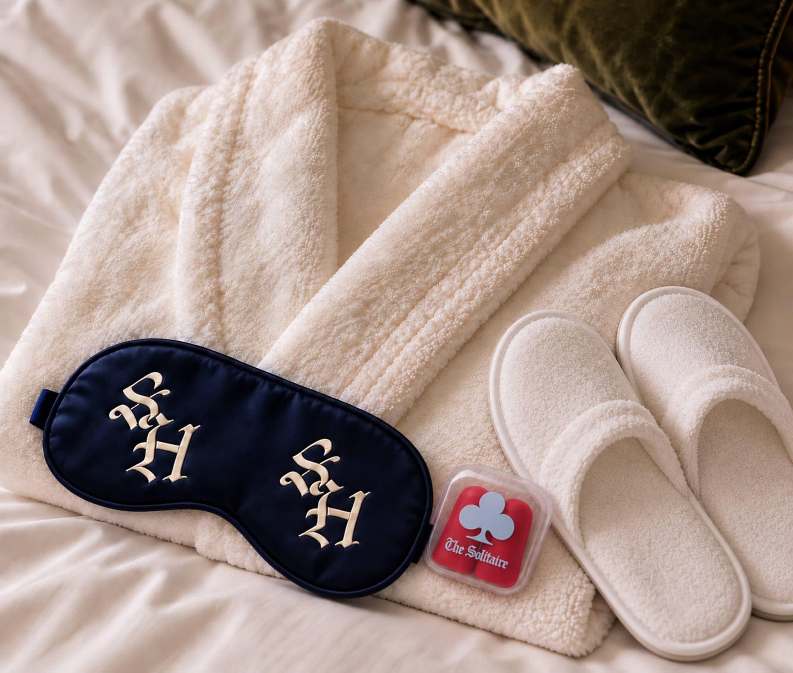 A neatly arranged set of white towels, a pair of slippers, and a sleep mask on a bed.