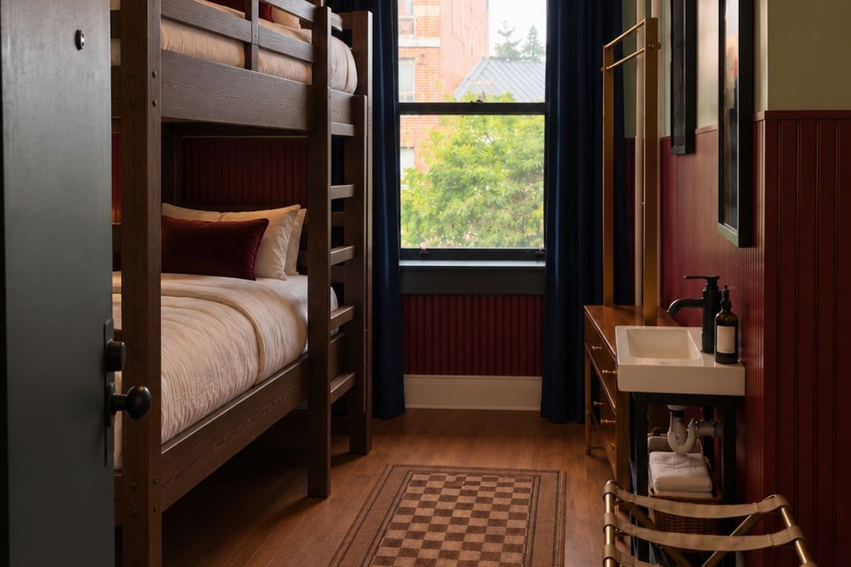 A room with a bunk bed, a desk with a sink, a chair, a window with curtains, and a hanging light fixture.