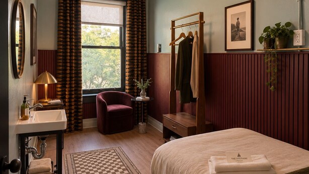 A hotel room with a checkered floor, a bed with a folded towel, a desk with a lamp, a chair, a window with curtains, and a wall-mounted coat rack. - The Solitaire Hotel (Vancouver)