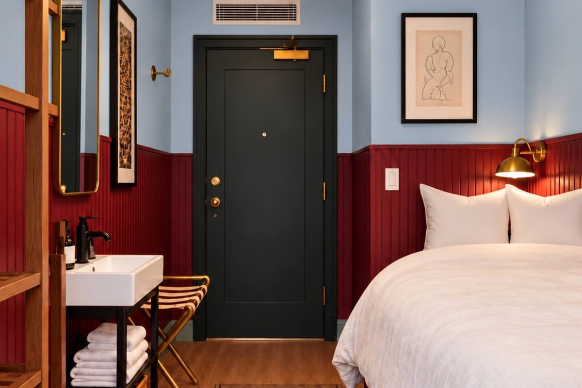 A hotel room with a bed, a bathroom with a sink and mirror, a door, and a wall-mounted light fixture.