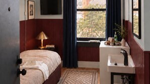 A hotel room with a bed, bedside lamp, window with curtains, a mounted TV, and a small sink with a mirror. - The Solitaire Hotel (Vancouver)