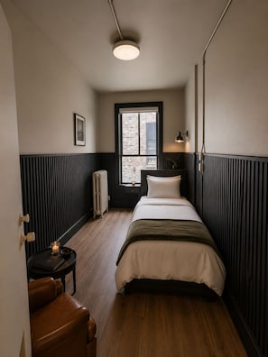 A bedroom with a bed, a window, a radiator, a bedside table, a lamp, and a chair. - The Solitaire Hotel (Vancouver)
