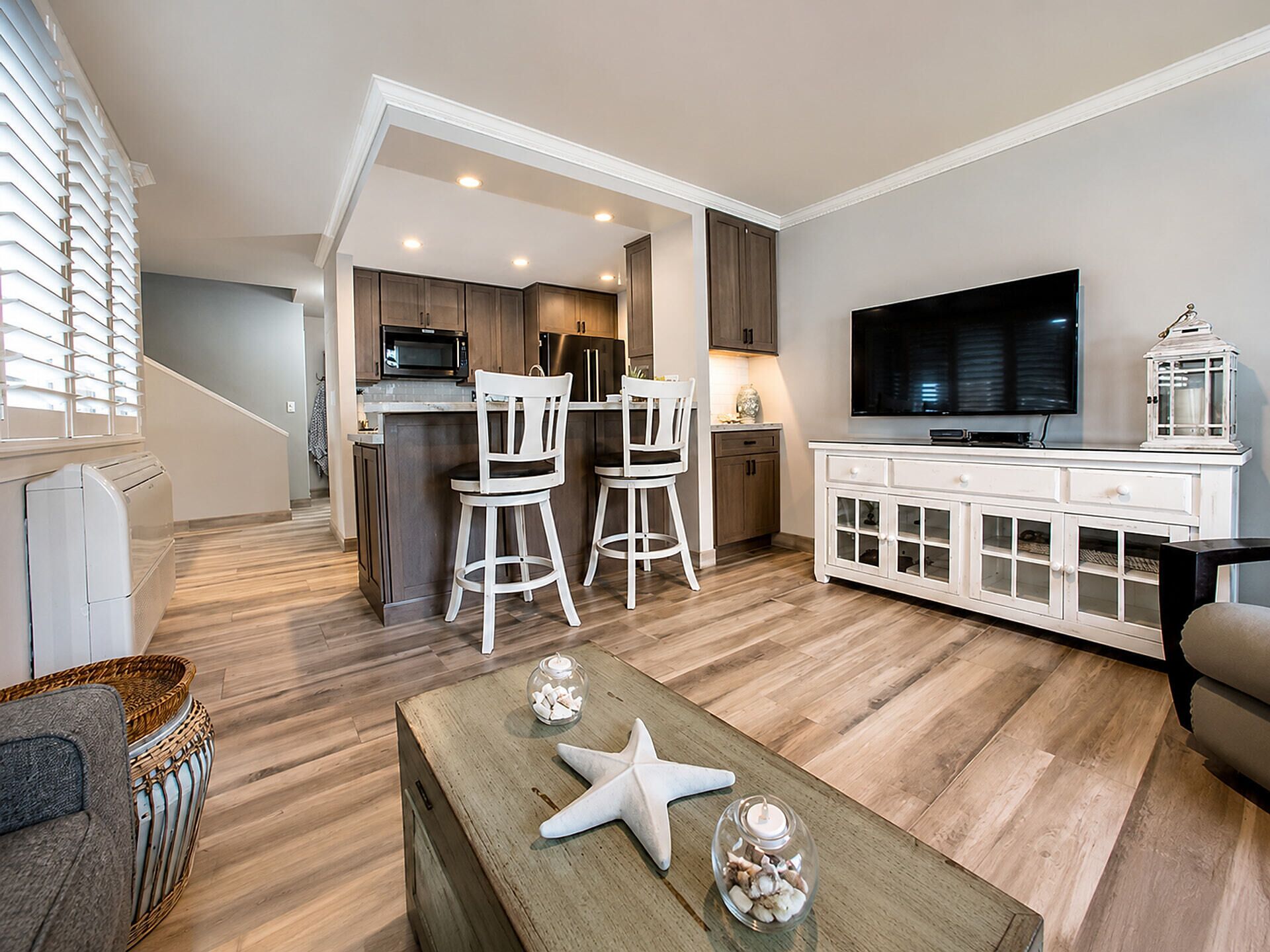 A modern living room with a wooden floor, a television, a white cabinet, and a kitchen area with dark cabinets and a microwave.