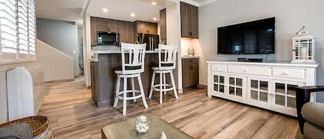 A modern living room with a wooden floor, a television, a white cabinet, and a kitchen area with dark cabinets and a microwave.