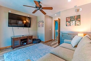 A living room with a ceiling fan, a television, a wooden entertainment unit, a couch, and a door leading to another room. - 🏖️ Luxury 8BR🌊 Ocean Views💦 Free Pool Heat♨️ Elevator🐾2nd Row✨Pet Friendly (Nagshead)