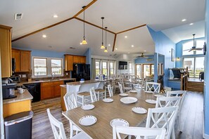 A spacious kitchen with wooden cabinets, a dining area with white chairs and plates, and a large window providing natural light. - 🏖️ Luxury 8BR🌊 Ocean Views💦 Free Pool Heat♨️ Elevator🐾2nd Row✨Pet Friendly (Nagshead)