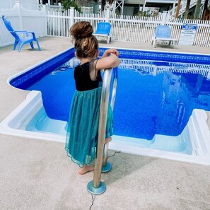 Un enfant se tient près d’une piscine, avec une clôture blanche et des chaises bleues en arrière-plan.