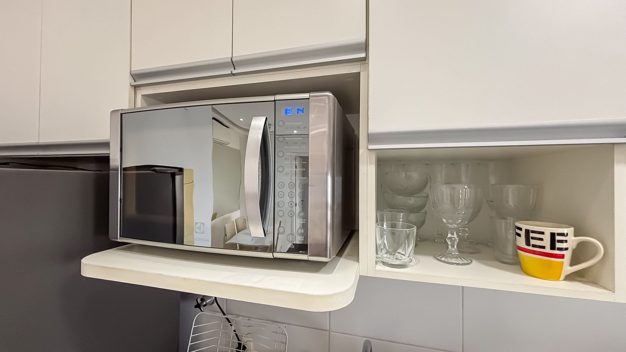 A kitchen with a microwave, a refrigerator, and a shelf with glasses and a mug.