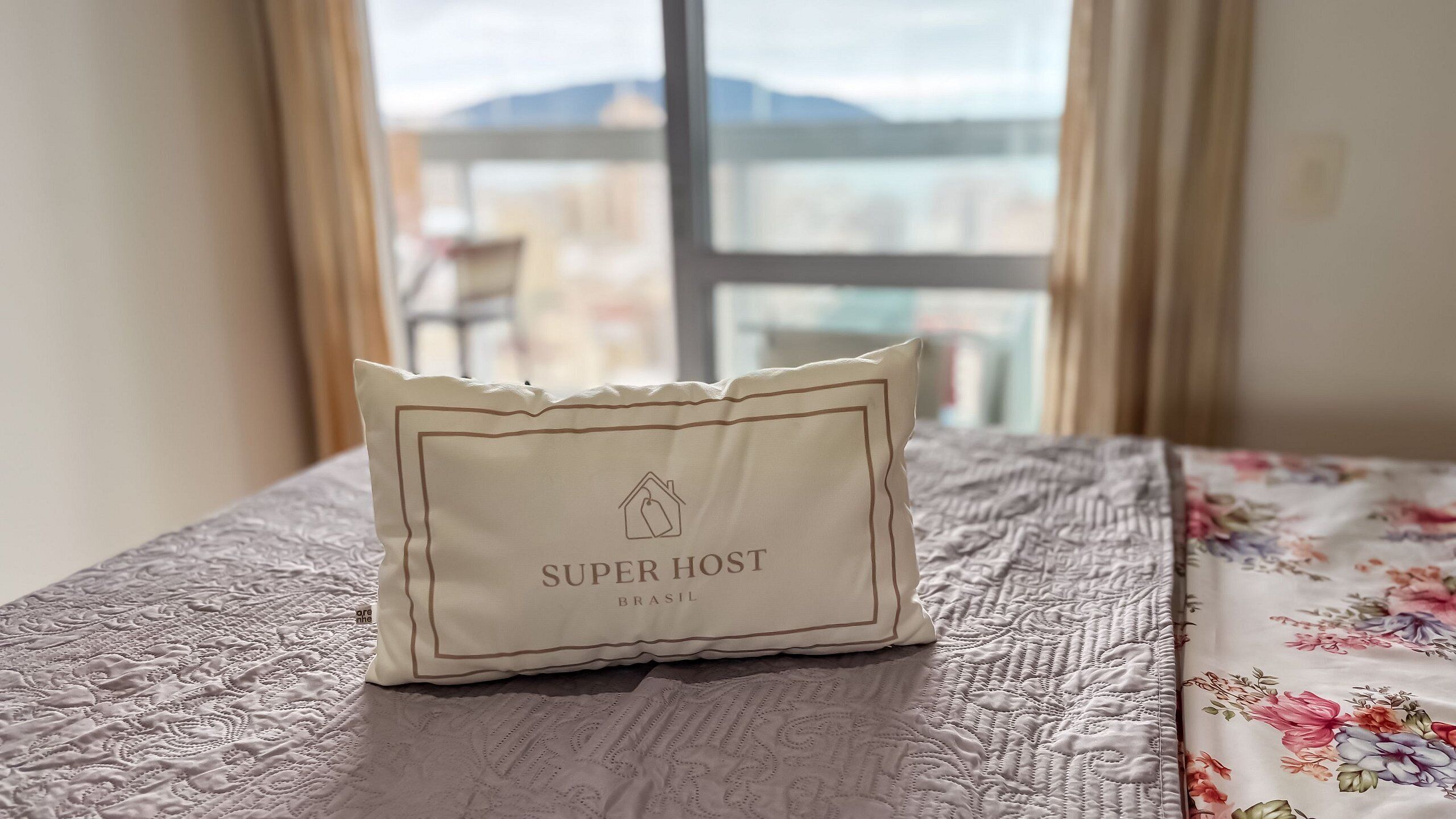 A pillow with 'SUPER HOST BRASIL' text on a bed with a floral bedspread.