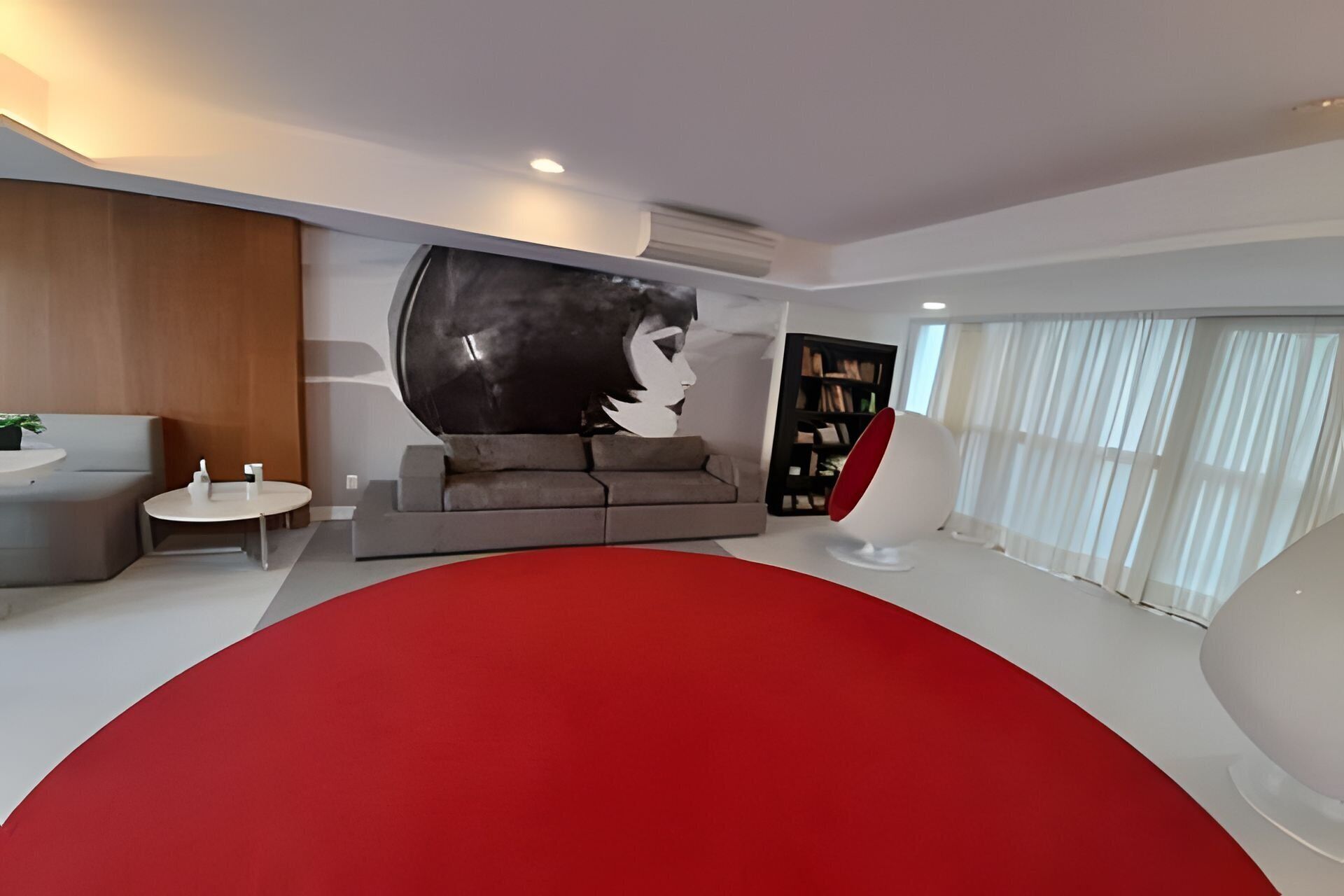 Un salón moderno con un tapete circular rojo grande, un sofá gris, una mesita redonda pequeña y una estantería.