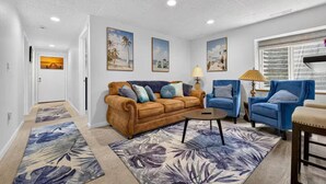 A living room with a brown sofa, blue armchairs, and a coffee table. There are three framed pictures on the wall, a lamp, and a window with blinds. - The little Beach House in the Forest (Colorado Springs)