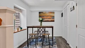 A hallway with a dining table, chairs, a vase with flowers, a painting on the wall, and a window with blinds. - The little Beach House in the Forest (Colorado Springs)