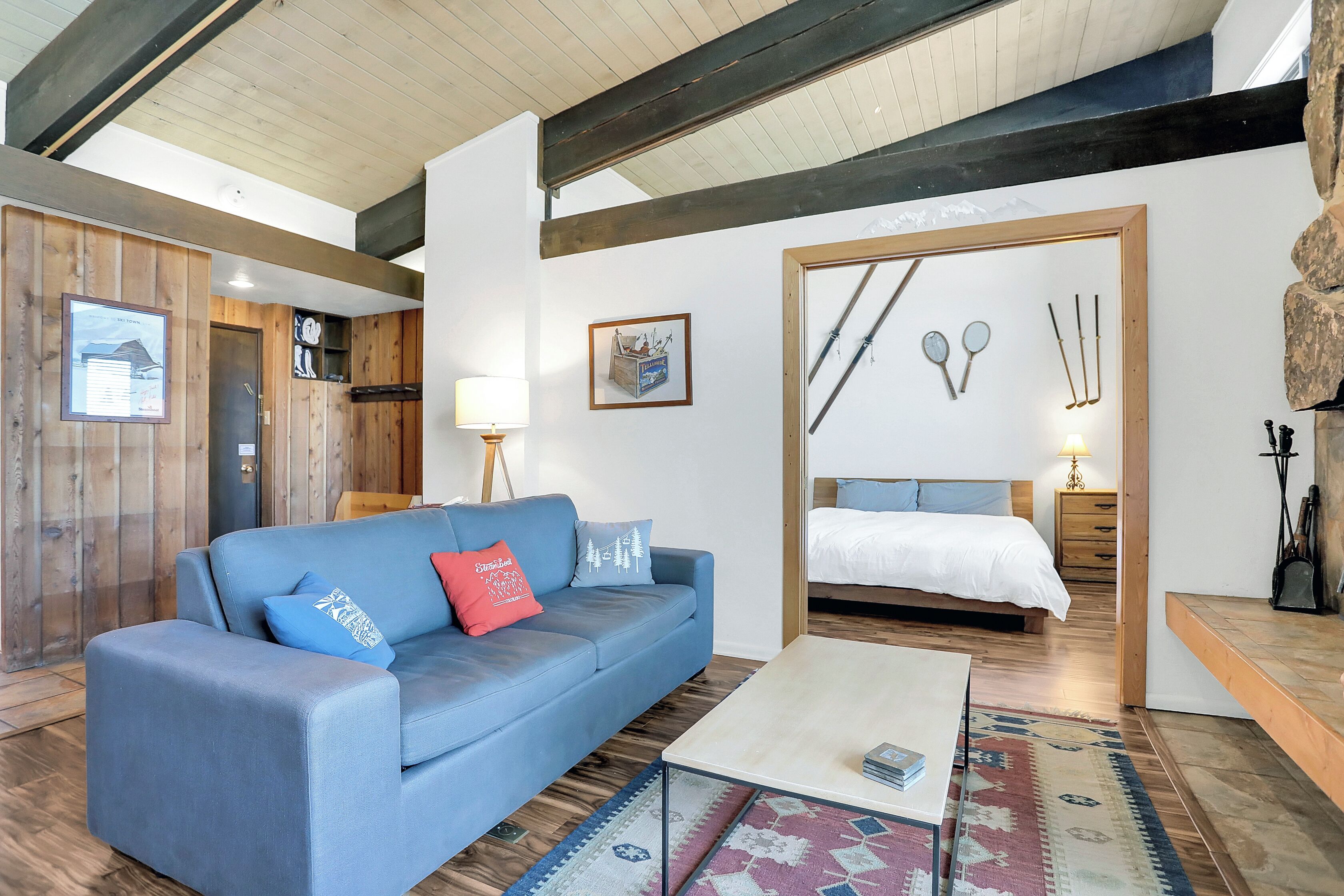 Un interior moderno con un sofá azul, piso de madera, cama con edredón blanco y decoración de pared con temática de esquí.