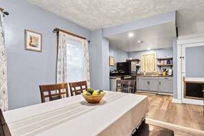 Un comedor con una mesa, sillas, un cuenco de fruta, un televisor y una cocina al fondo.