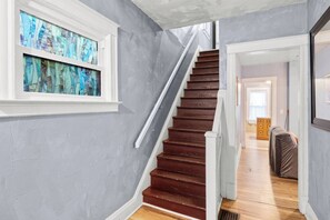 A staircase with wooden steps and a white handrail, leading to a room with a gray wall and a window with stained glass. - 2 Homes • Sleeps 12 • 6BR • 1 Mile to Falls (Niagara Falls)