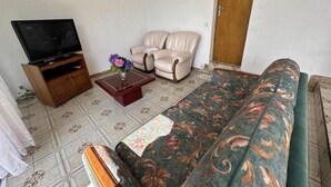 Ein Wohnzimmer mit einer Couch, zwei Sesseln, einem Couchtisch mit einer Vase, einem Fernseher auf einem Holzständer und einem gemusterten Teppich.
