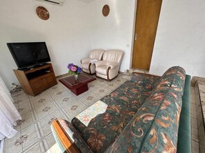 Ein Wohnzimmer mit einer Couch, zwei Sesseln, einem Couchtisch mit einer Vase, einem Fernseher auf einem Holzständer und einem gemusterten Teppich.